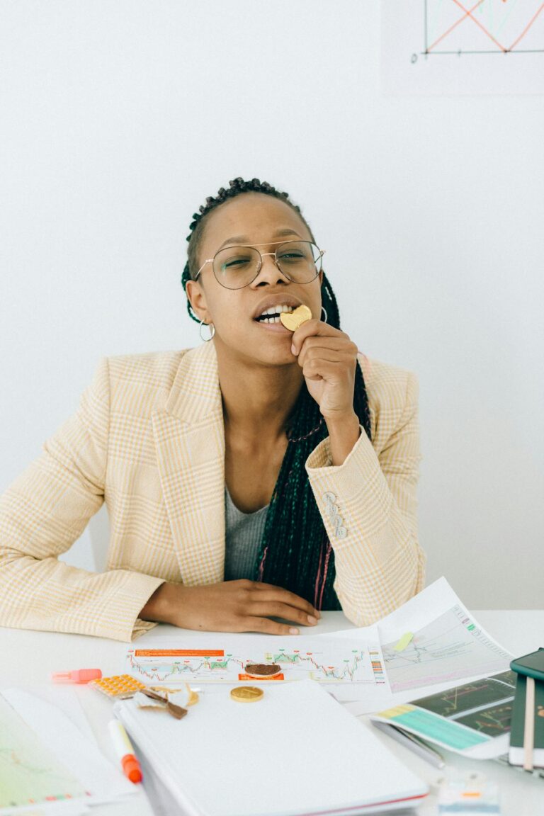 A professional woman examines financial documents and bites a chocolate coin at a white desk indoors.